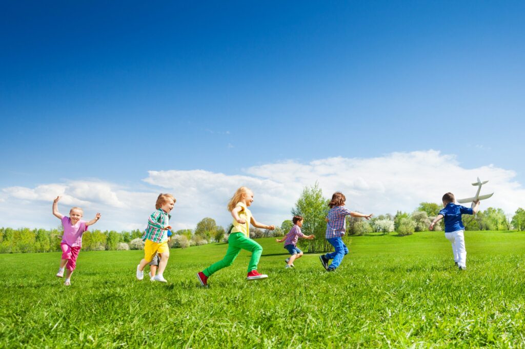 A group of children goes to play with a toy airplane on a grassy field under a clear blue sky, surrounded by a lightly wooded area.