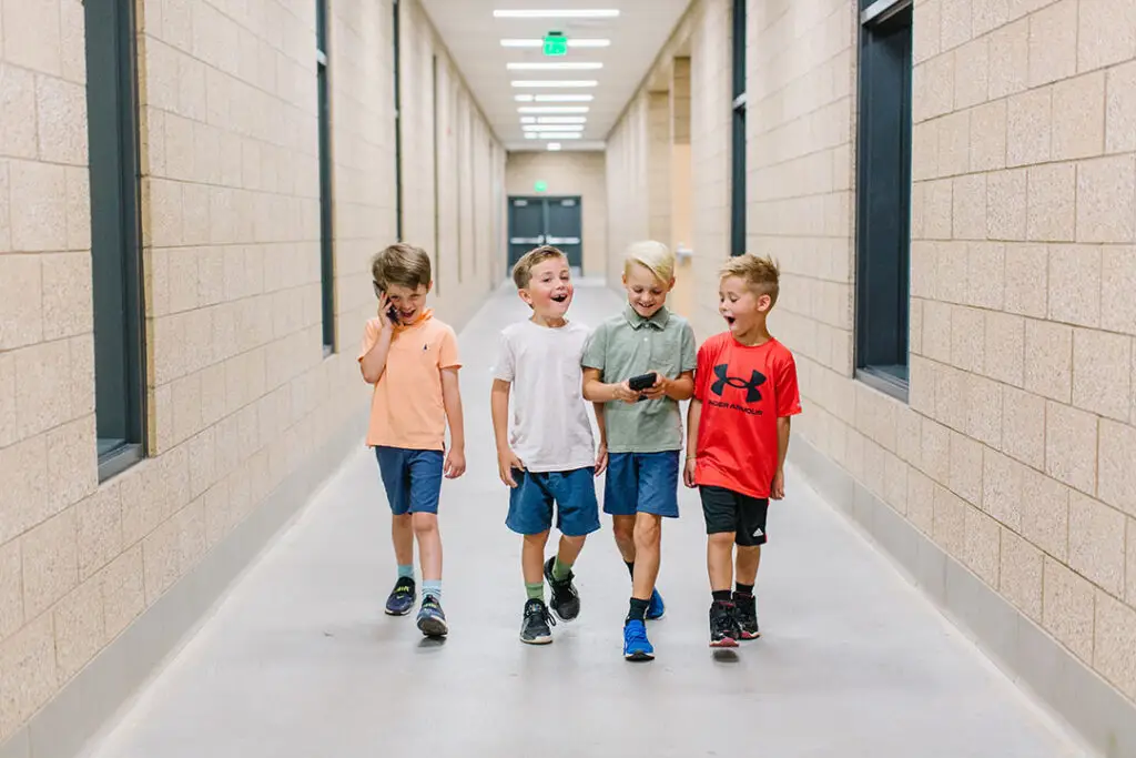 Four children stroll down a school hallway, smiling and chatting. One holds a cell phone, and they look casual in their summer clothes. The corridor stretches long with beige brick walls and doors lining the sides.