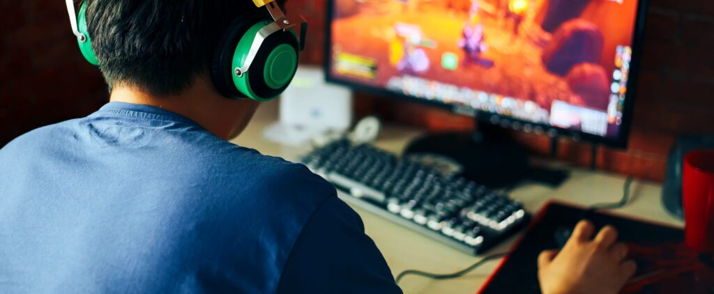 A person wearing green headphones plays a video game on the computer, immersed in a colorful fantasy interface. With a keyboard and mouse on the desk, their focus is intense, navigating through thrilling quests that blur the lines between reality and gambling in video games.