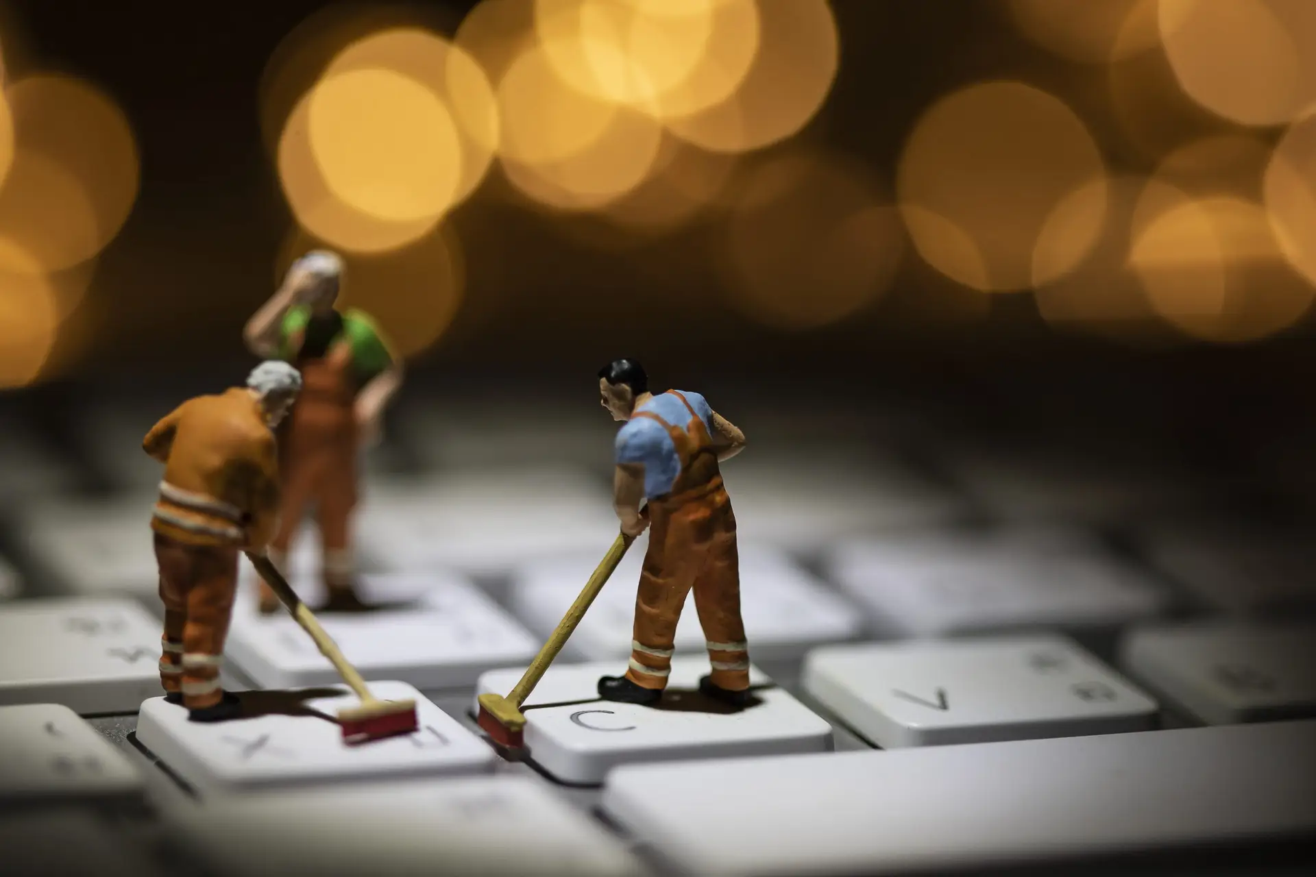 Three miniature figures in work attire diligently sweep a computer keyboard with brooms, embodying the question: How can we keep our technology clean? The scene is artistically lit against a warm, blurry background of circular lights.