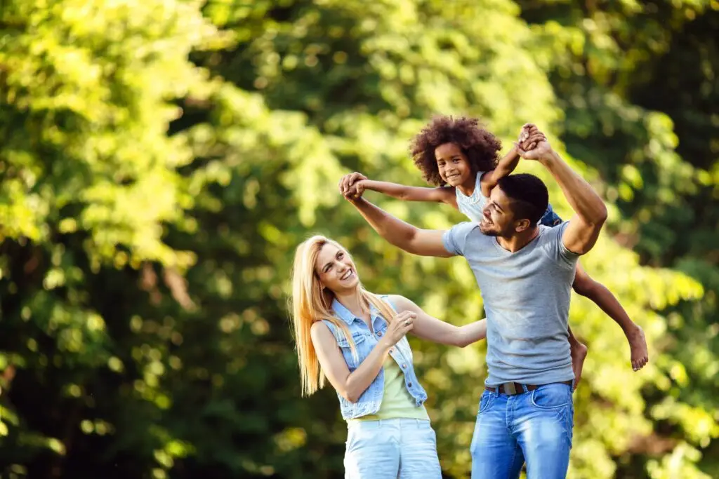 A joyful family enjoys the benefits of going outside on a sunny day in a park. A man playfully lifts a child by the arms while a woman smiles beside them. The background features lush green trees, creating a vibrant, cheerful atmosphere.