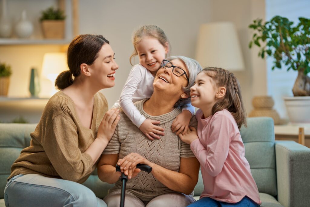 A joyful elderly woman, seated and holding a walking stick, is surrounded by two young girls and a woman. They are embracing and smiling in a cozy living room with plants and soft lighting, embodying the warmth of intensive parenting in a loving atmosphere.