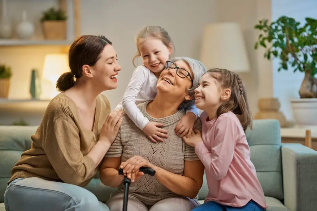 A joyful elderly woman, seated and holding a walking stick, is surrounded by two young girls and a woman. They are embracing and smiling in a cozy living room with plants and soft lighting, embodying the warmth of intensive parenting in a loving atmosphere.