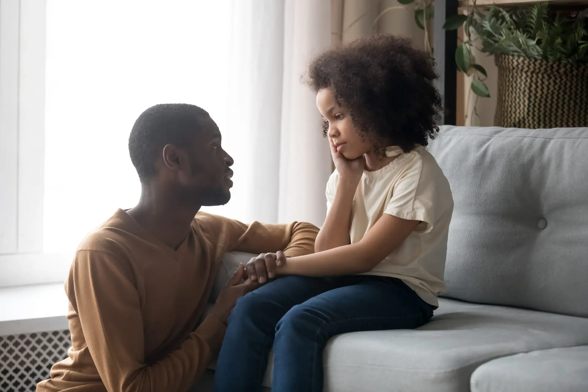 A man kneels beside a seated child on a couch, holding hands and facing each other as the adult gently discusses emotional regulation for kids. The child appears thoughtful, resting their chin on one hand. The setting is a cozy room with soft lighting and plants.