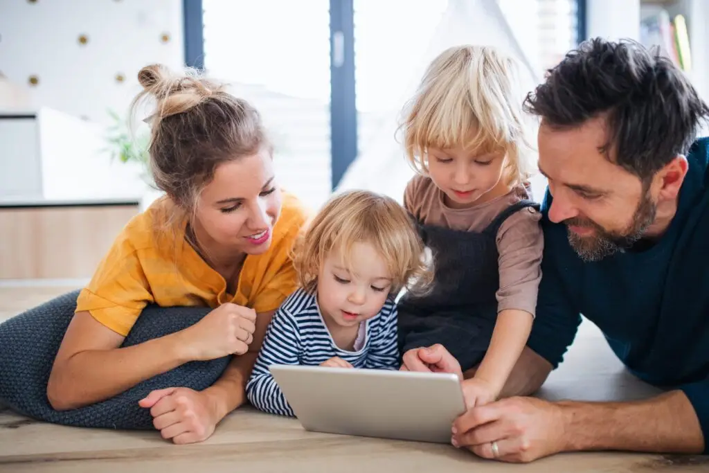 A family of four, including two young children, is lying on the floor at home, happily exploring a tablet together. The cozy and bright environment adds to their joy as they delve into fun topics like "What is a Firewall and How Does it Work?