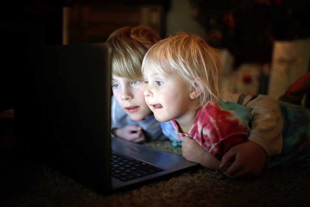 Two children lie on the floor, closely watching a laptop screen, its soft glow illuminating their focused expressions. The older child with light brown hair and the younger blond, ponder silently in the dimly lit, cozy background—a curious moment that might lead them to wonder: is the Dark Web safe?