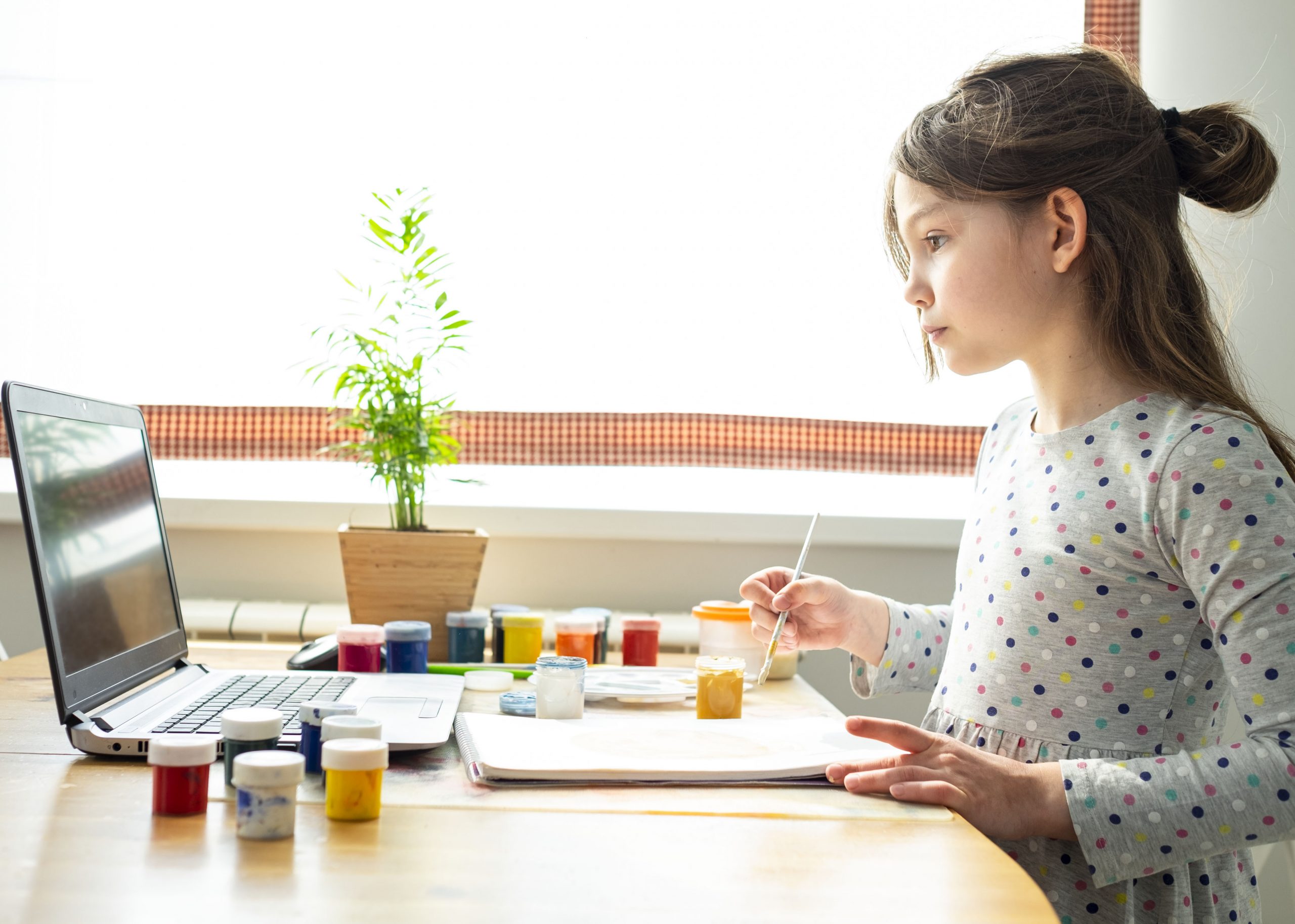 A young girl in a polka-dot shirt paints at a table with an open laptop nearby, perhaps exploring apps for art inspiration. Various paint jars are scattered around, and a small potted plant is in the background. Sunlight streams in through a window, illuminating the scene.