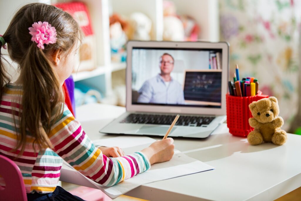 A young girl with a floral hair clip studies at her desk, writing in a notebook. A laptop before her displays a teacher, thanks to online learning platforms for kids. Her desk is dotted with colorful pens and a teddy bear companion.