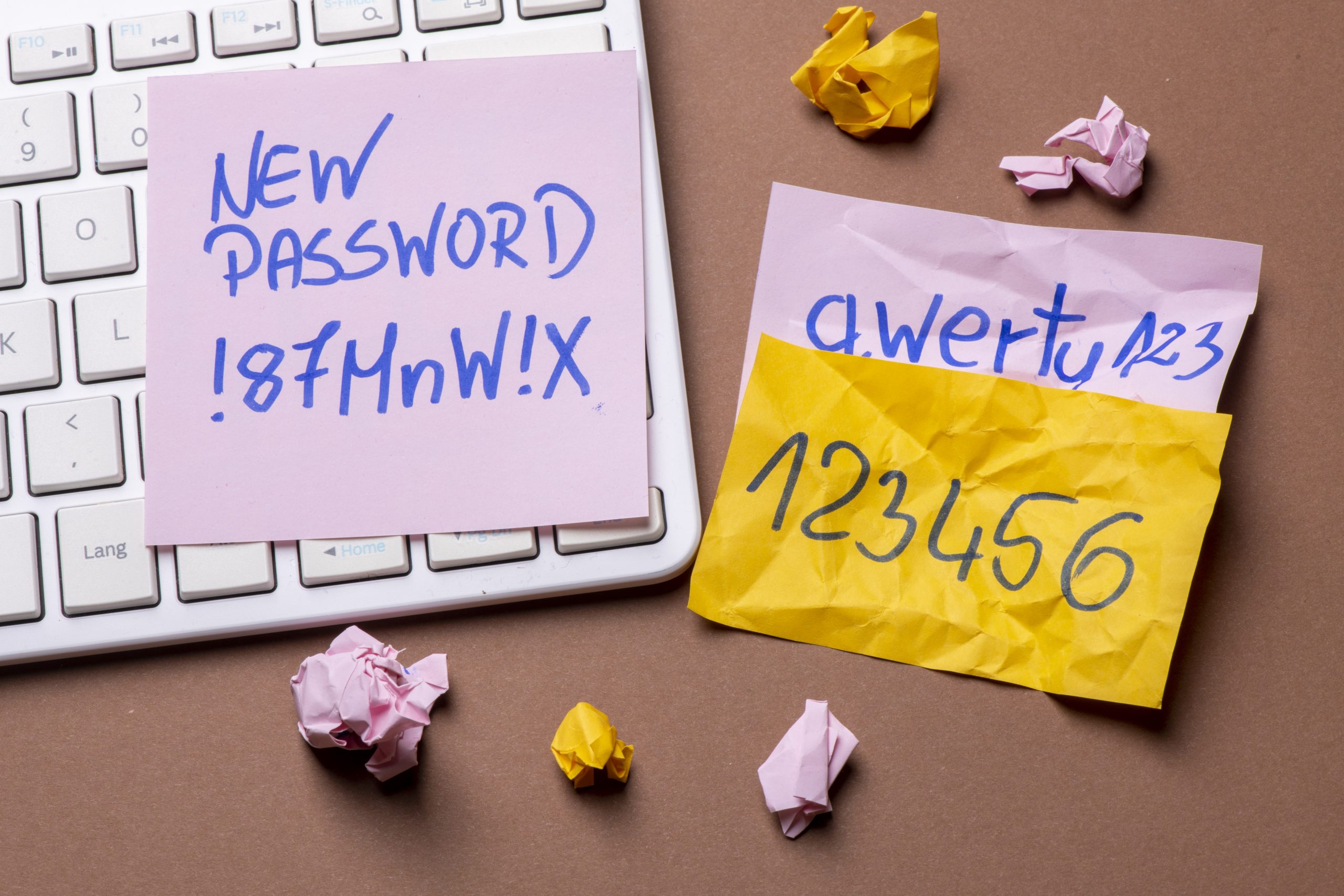 A keyboard cluttered with sticky notes reveals the importance of strong passwords—handwritten reminders like New Password !87MnW!X on a pink note and a.wertu123 with 123456 on yellow. Crumpled papers scattered around underscore the chaos of weak security measures.