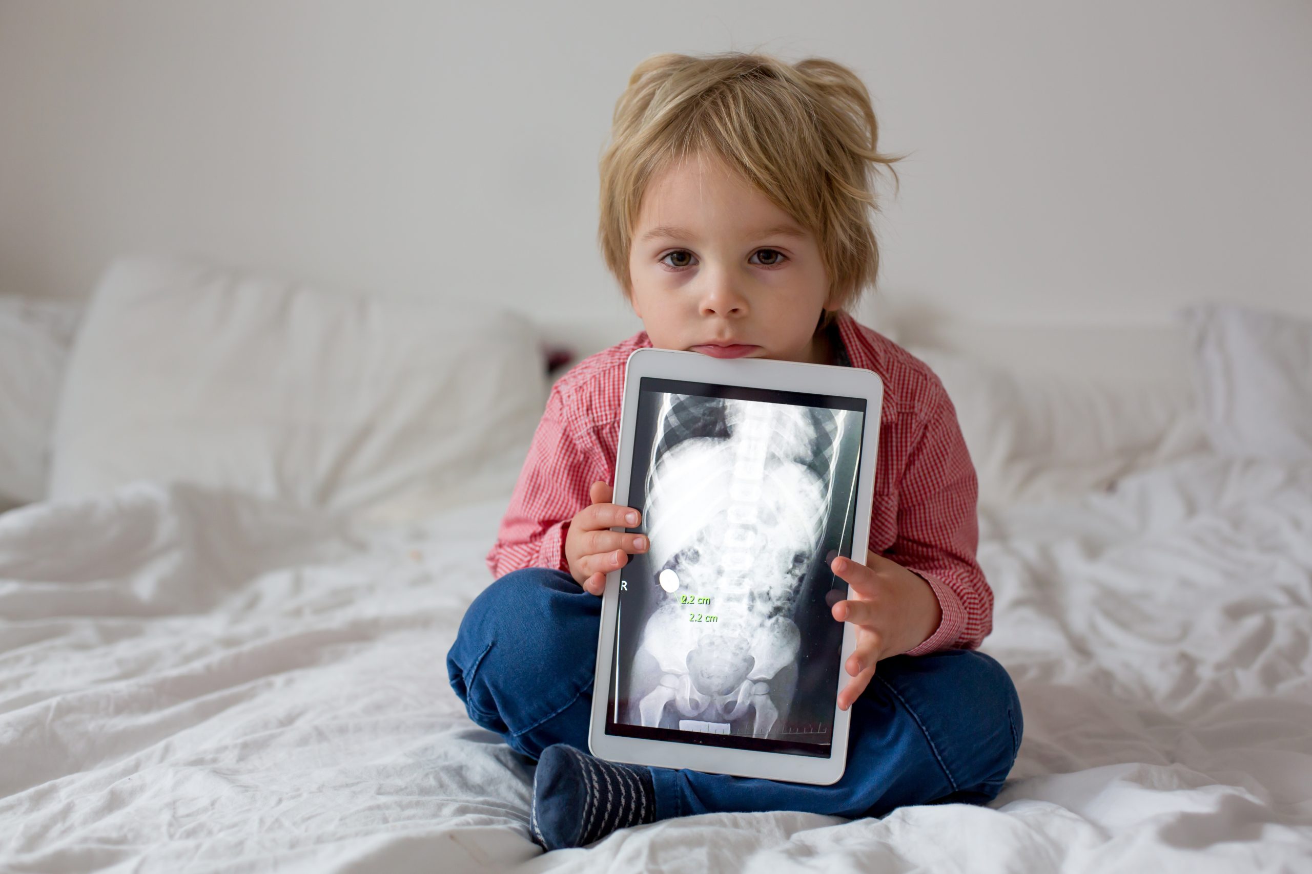 A young child with blond hair sits on a bed, holding a tablet displaying an X-ray image. Wearing a red shirt and blue jeans, the child looks attentively at the camera, possibly learning about how high-powered magnets can harm children. The background is a plain white wall.