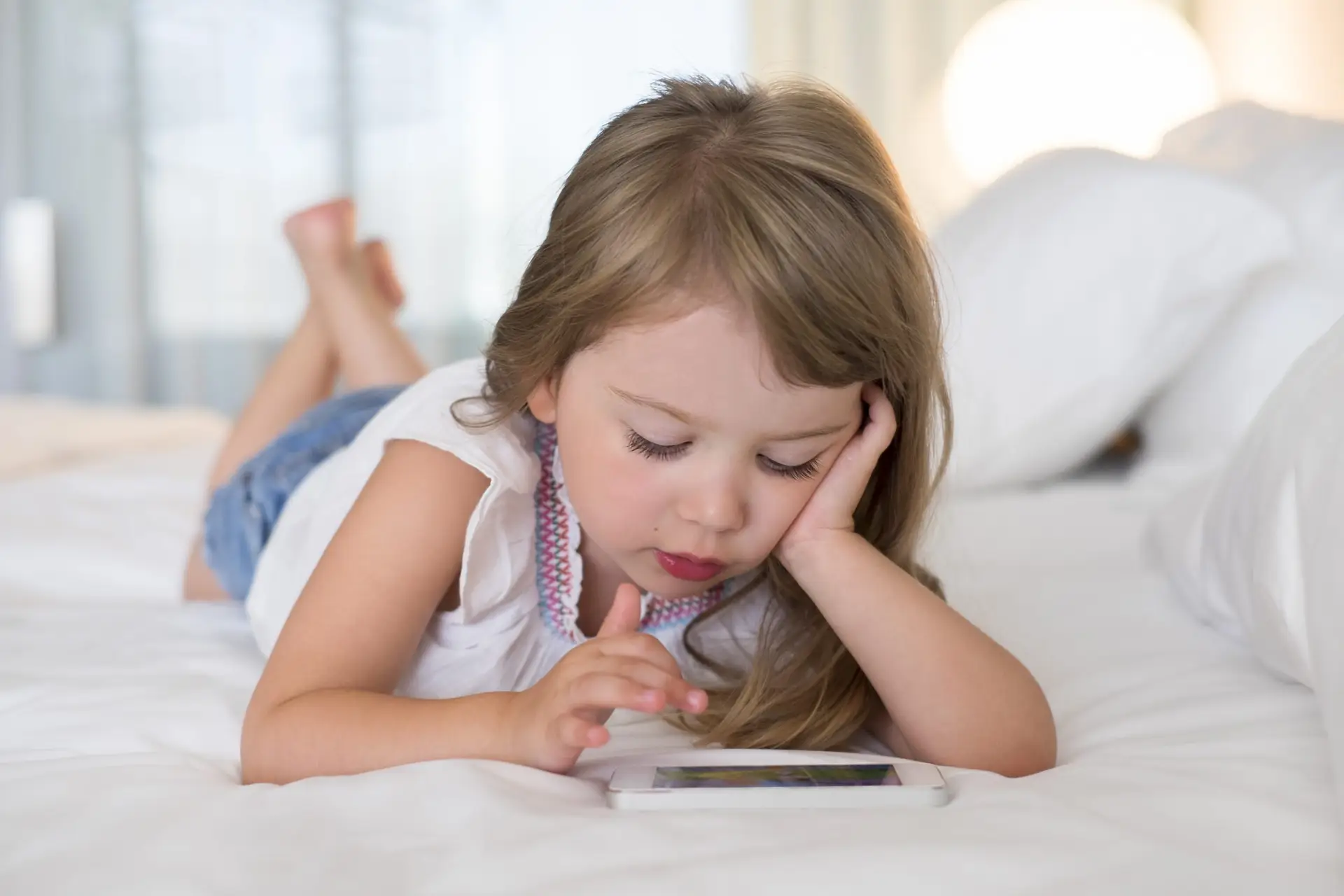 A young girl with long brown hair is lying on a bed, seemingly lost in too much screen time. She wears a white sleeveless top and denim shorts, her head resting on one hand while the other taps away at the smartphone's screen.