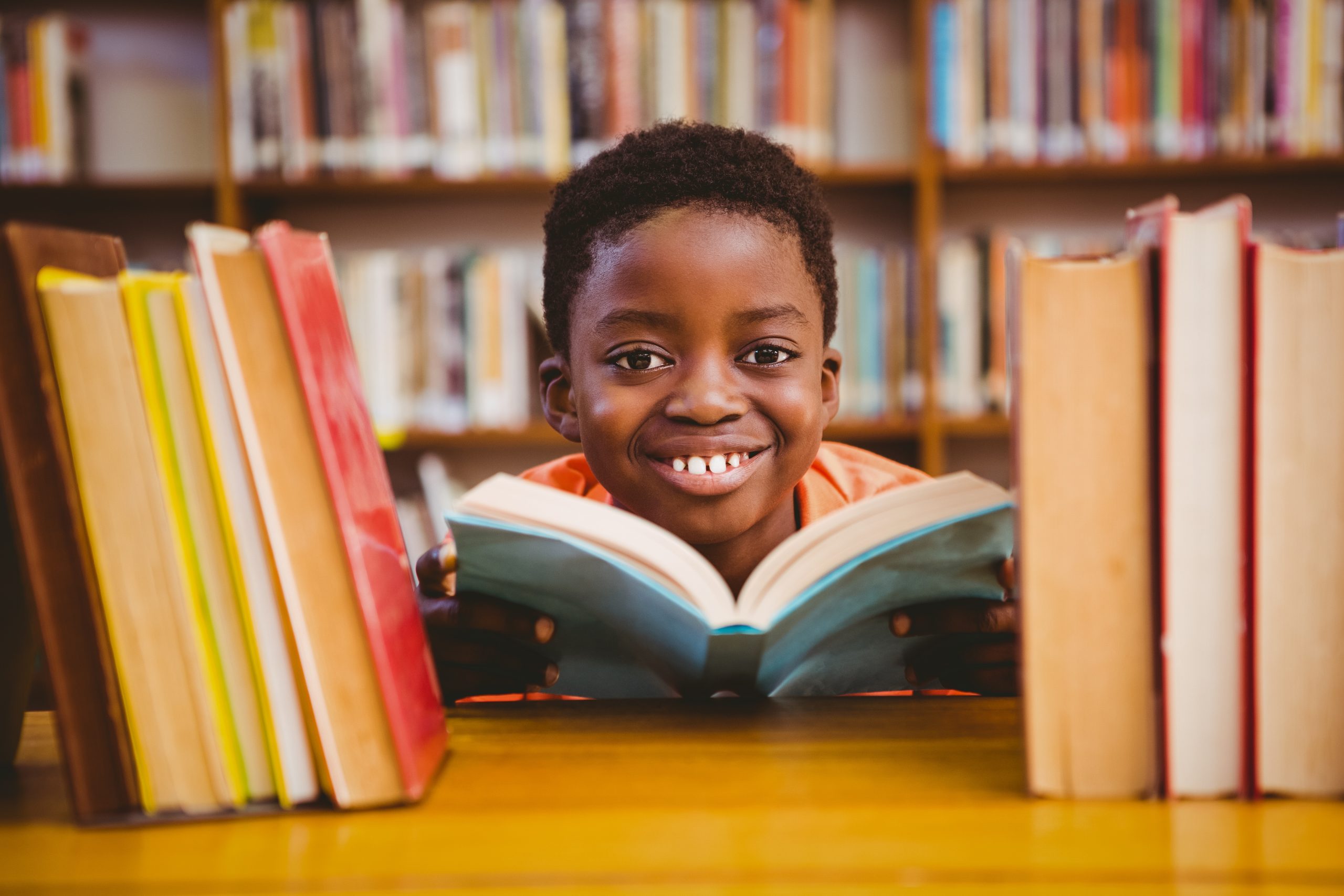 A smiling child is enjoying the benefits of reading for kids, nestled in a library surrounded by shelves of colorful books. The slightly blurred background highlights the joy of diving into new stories and adventures.