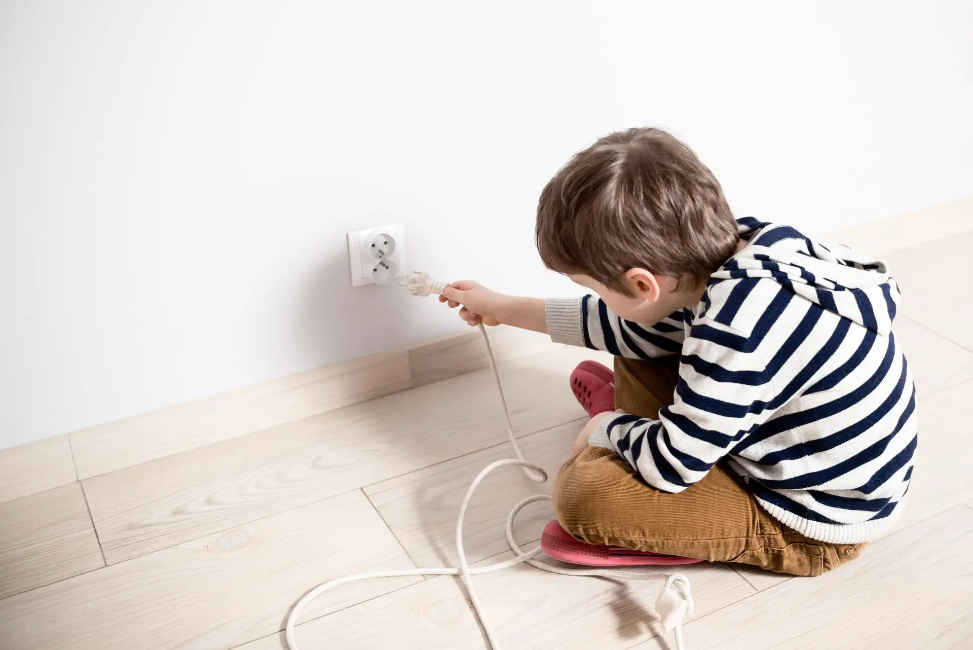 A child with brown hair, in a striped hoodie and brown pants, sits on a wooden floor. Practicing toddler safety, they gently hold a plug near a wall socket, with a long cable coiled beside them.