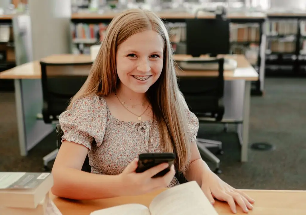 A young girl with long hair and braces smiles while holding a smartphone, highlighting technology as a learning tool. She's seated at a table in a library, an open book in front of her and shelves of books in the background.