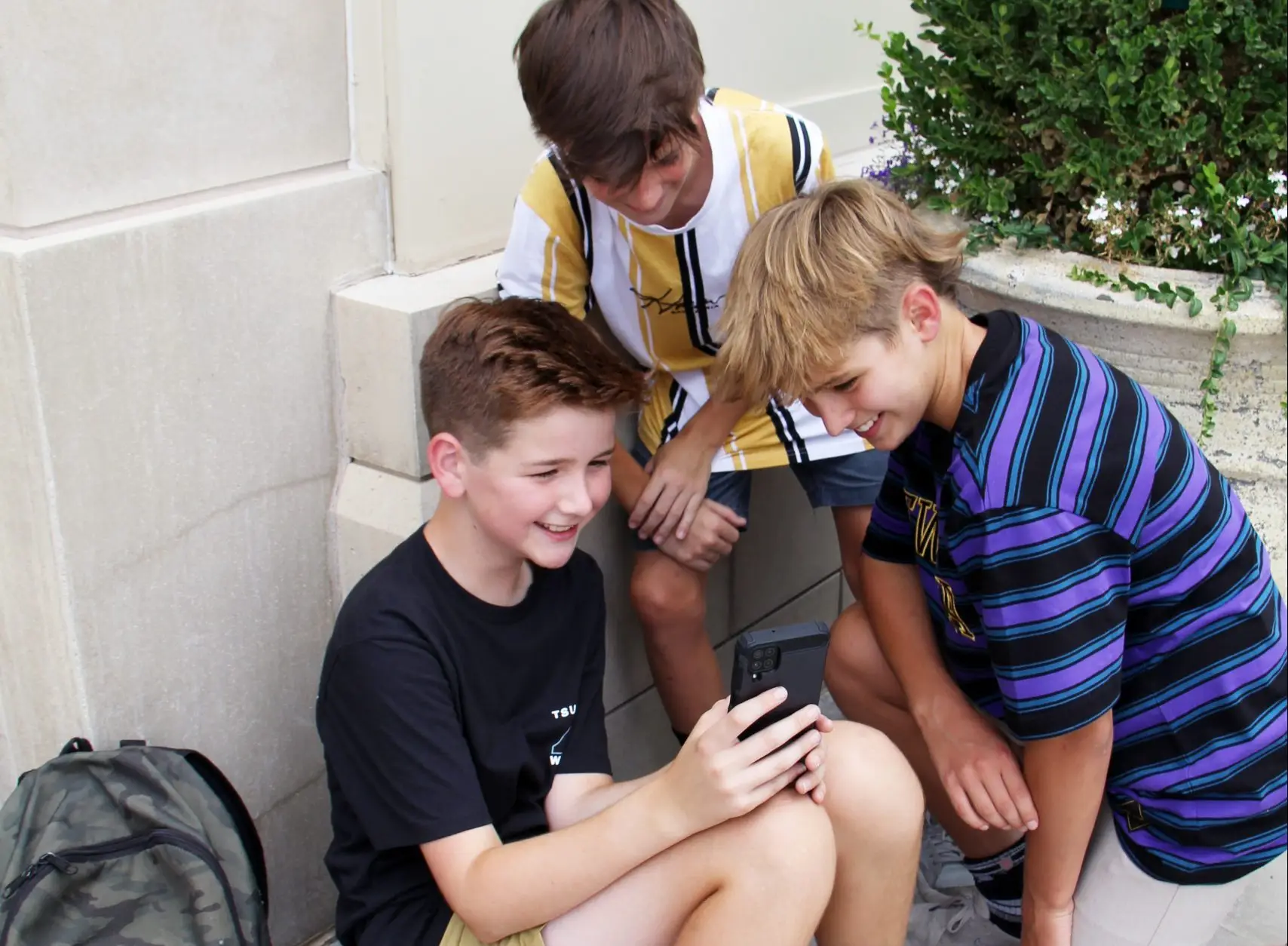 Three boys are huddled together outside, smiling as they explore healthy ways to use technology. One sits holding the smartphone, while the other two crouch beside him, engrossed. A backpack rests nearby amidst lush plants in the background.