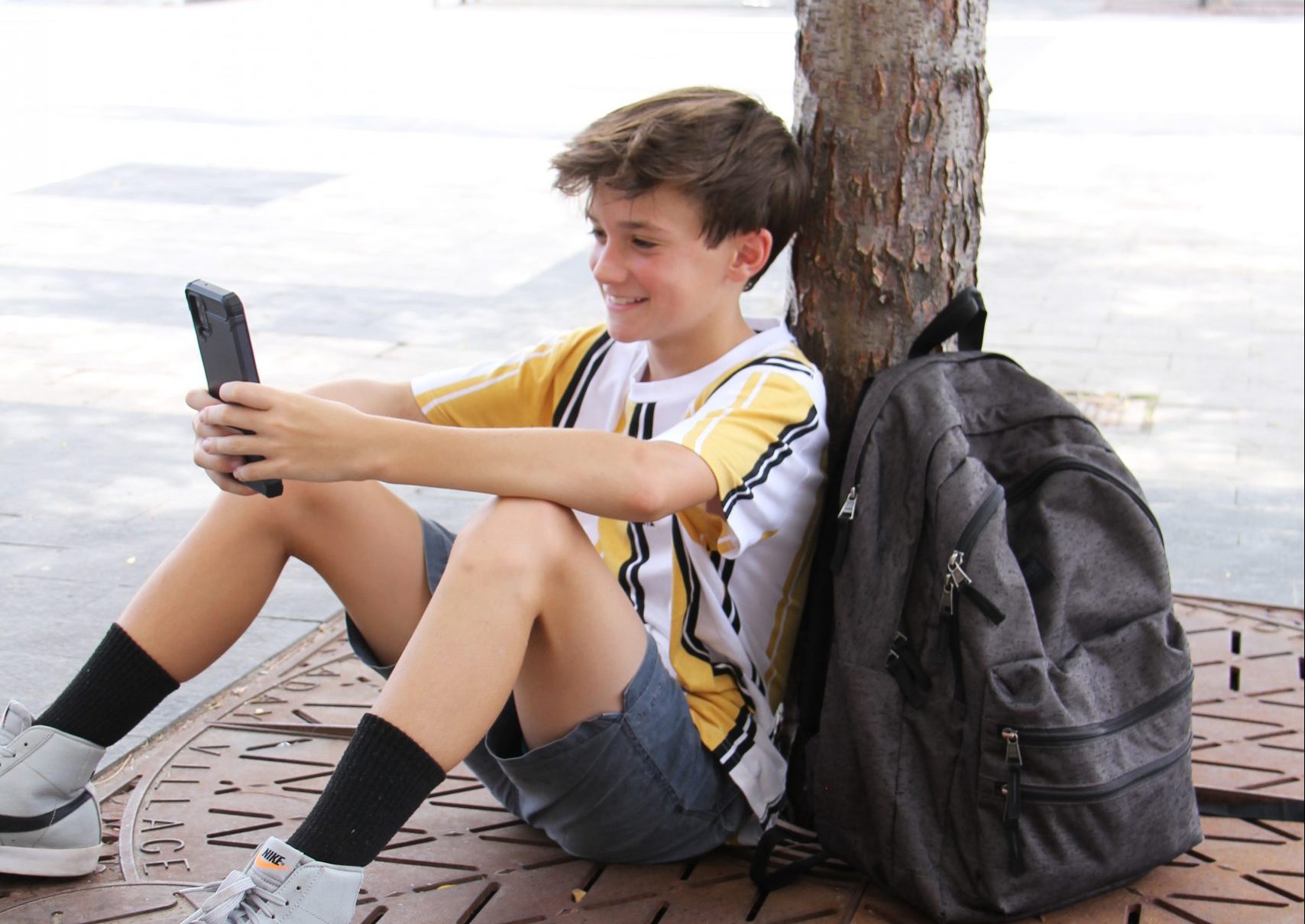 A boy in a yellow and white striped shirt sits under a tree, leaning against the trunk, smiling at a smartphone. Conscious of child privacy online, he's enjoying his screen time responsibly. A gray backpack rests beside him in this outdoor urban area.