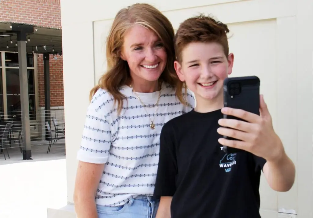 A woman and a young boy are smiling as they take a selfie outdoors. The woman is wearing a striped shirt, while the boy, holding a smartphone with enabled notifications, captures the happy and relaxed moment perfectly.