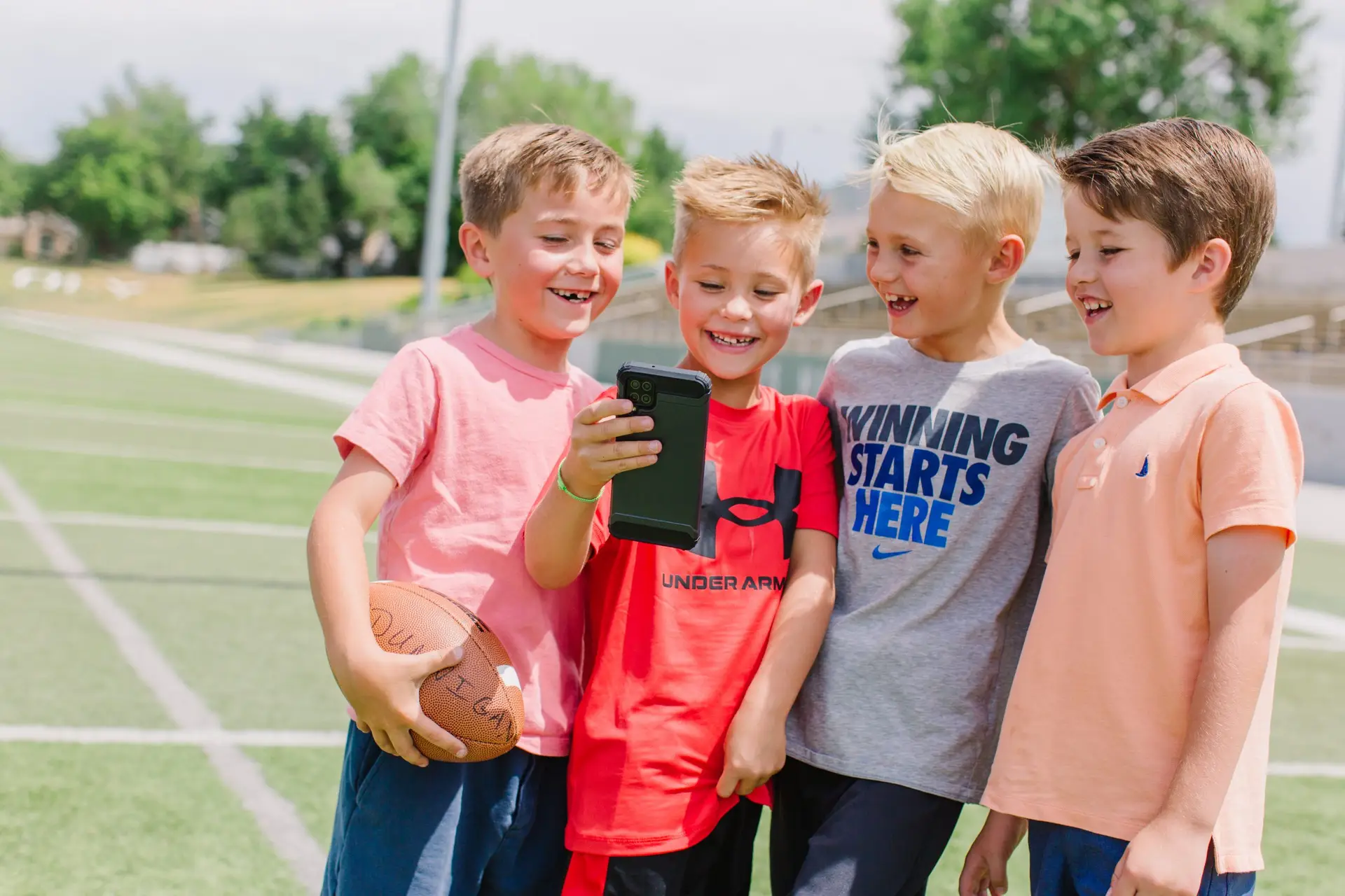 Four young boys stand on a football field, smiling as they huddle around a smartphone held by one of them. With the sun shining and yard lines clearly marked, one boy grips a football, sparking the question—when should kids have a phone?