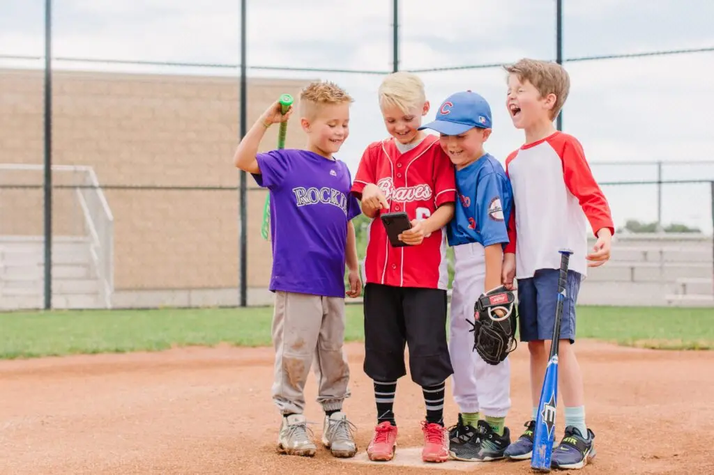 Four children in baseball uniforms stand on a baseball field, smiling and looking at a phone. They wear jerseys from different teams, embracing the joy of why kids should play sports. One holds a bat, with a chain-link fence and empty bleachers in the background.