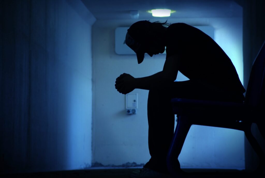 Silhouette of a person sitting on a bench in a dimly lit hallway, head bowed and hands clasped, evoking the contemplative atmosphere of the "Hear Me Out: Suicide Prevention" campaign.