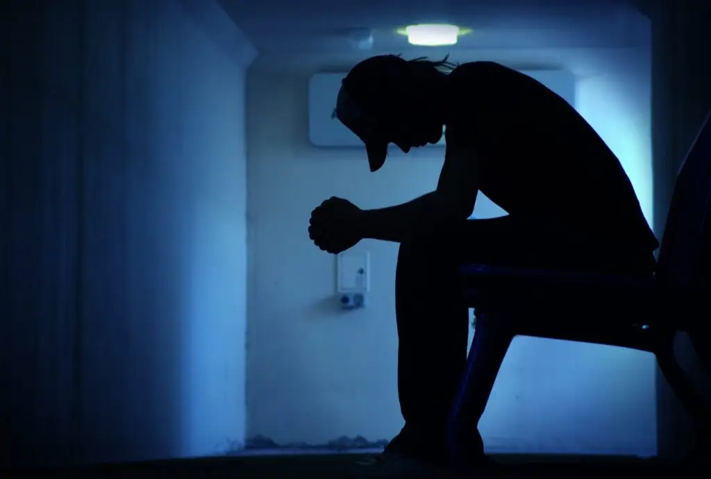 Silhouette of a person sitting on a bench in a dimly lit hallway, head bowed and hands clasped, evoking the contemplative atmosphere of the "Hear Me Out: Suicide Prevention" campaign.