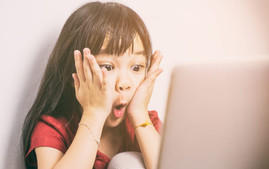 A young girl with long dark hair wearing a red shirt is sitting in front of a laptop, holding her face with both hands and displaying a surprised expression. Bright light illuminates her from the right side, highlighting the importance of COPPA Law in safeguarding children's privacy online.