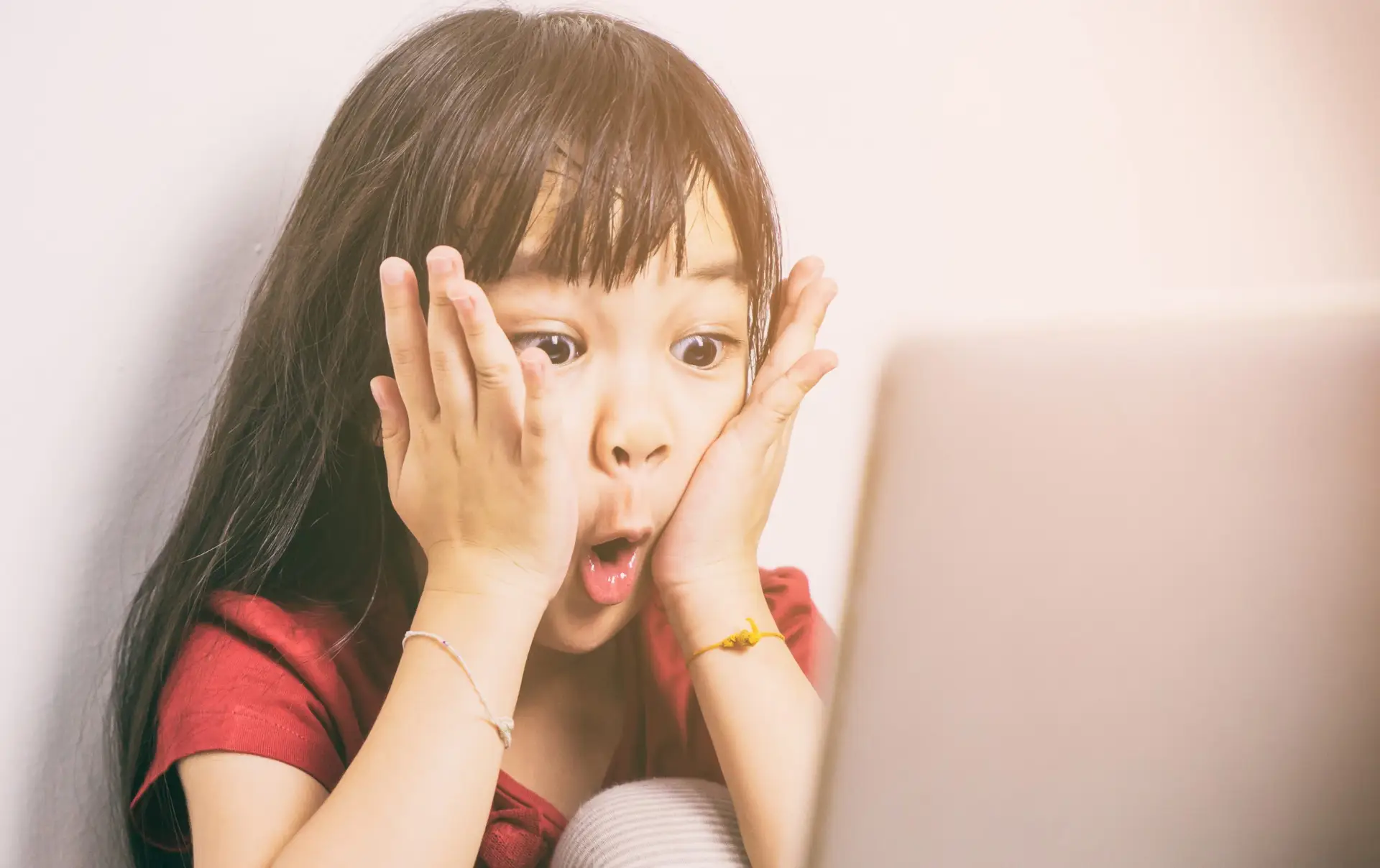 A young girl with long dark hair wearing a red shirt is sitting in front of a laptop, holding her face with both hands and displaying a surprised expression. Bright light illuminates her from the right side, highlighting the importance of COPPA Law in safeguarding children's privacy online.