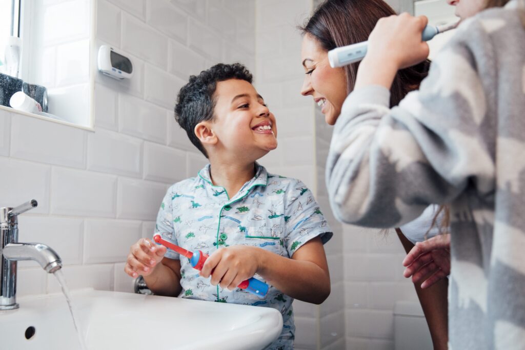 A young boy in a patterned shirt smiles while holding a red toothbrush by the sink, showcasing the benefits of routines. He is with two adults, one brushing their teeth with an electric toothbrush. The bathroom features white tiled walls and a small window to the left.