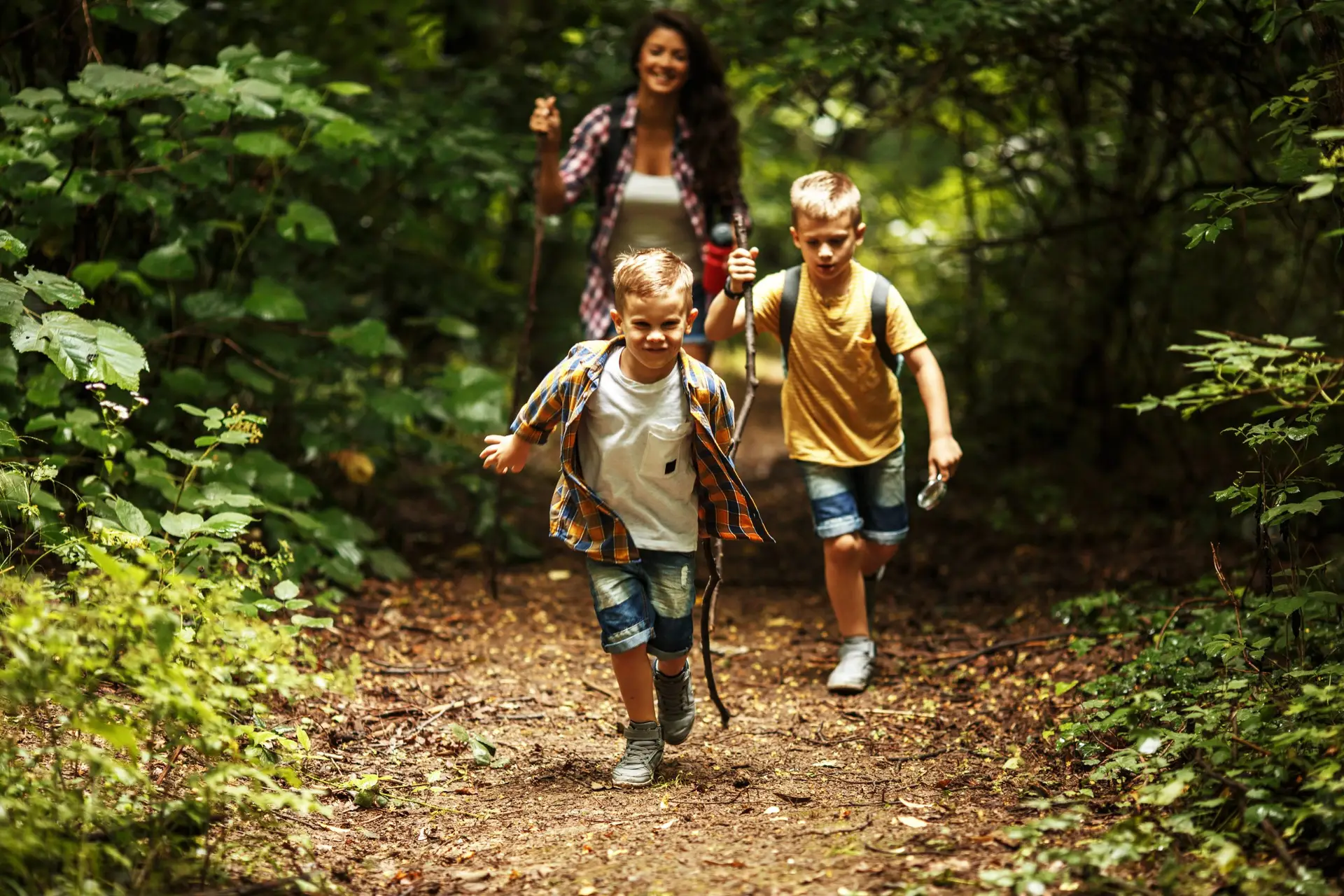 Two young boys happily run along a forest path, holding sticks, while an adult woman follows behind them. With lush green trees providing a vibrant backdrop, they embody one of the six simple tips for hiking with kids: keep the adventure light-hearted and joyful. All enjoy their day outdoors.