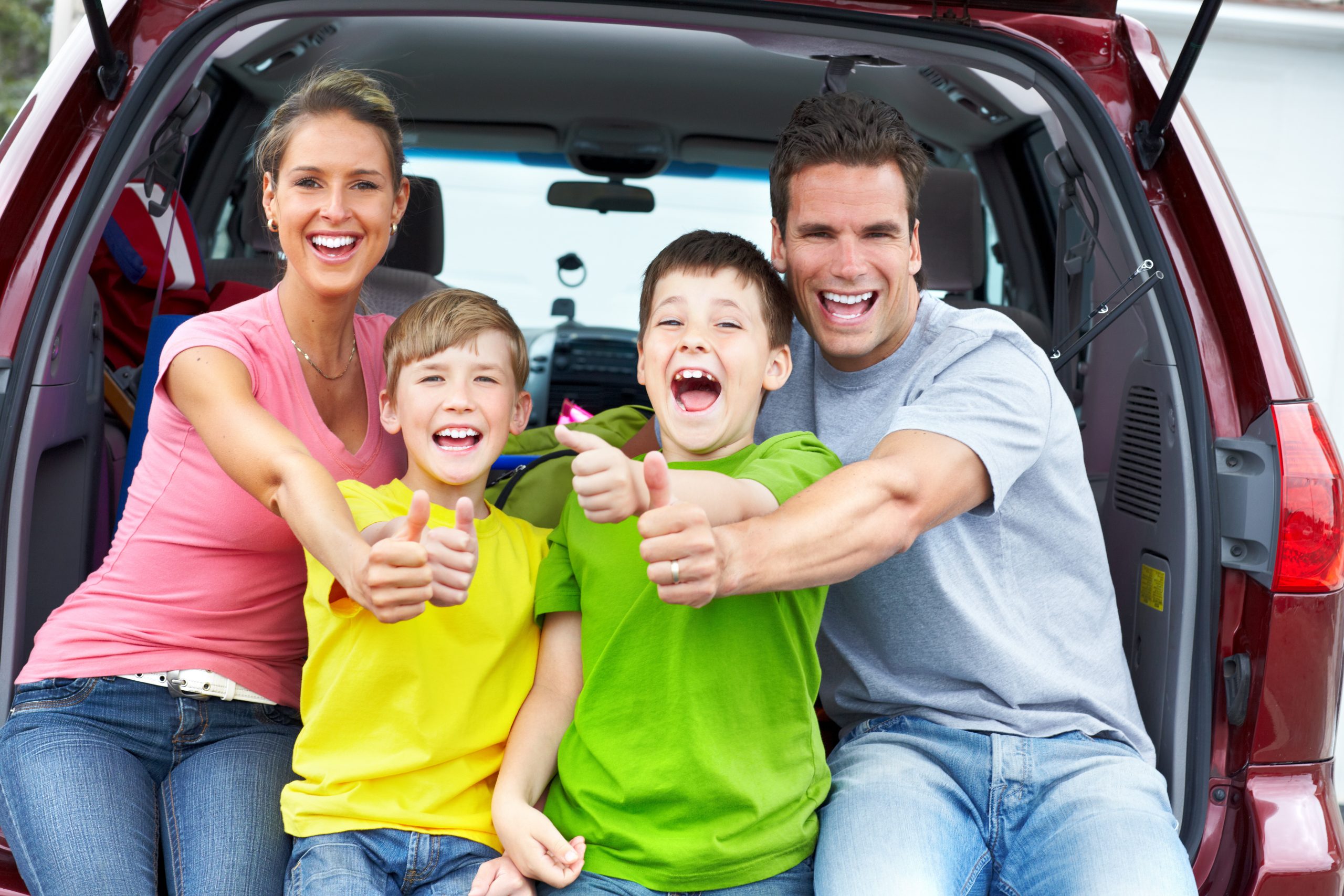 A smiling family of four sits in the back of an open car trunk. The parents, wearing casual clothes, sit at the ends while two young boys in yellow and green shirts occupy the middle. All show thumbs up, savoring a joyful moment together during their road trip activities for kids.