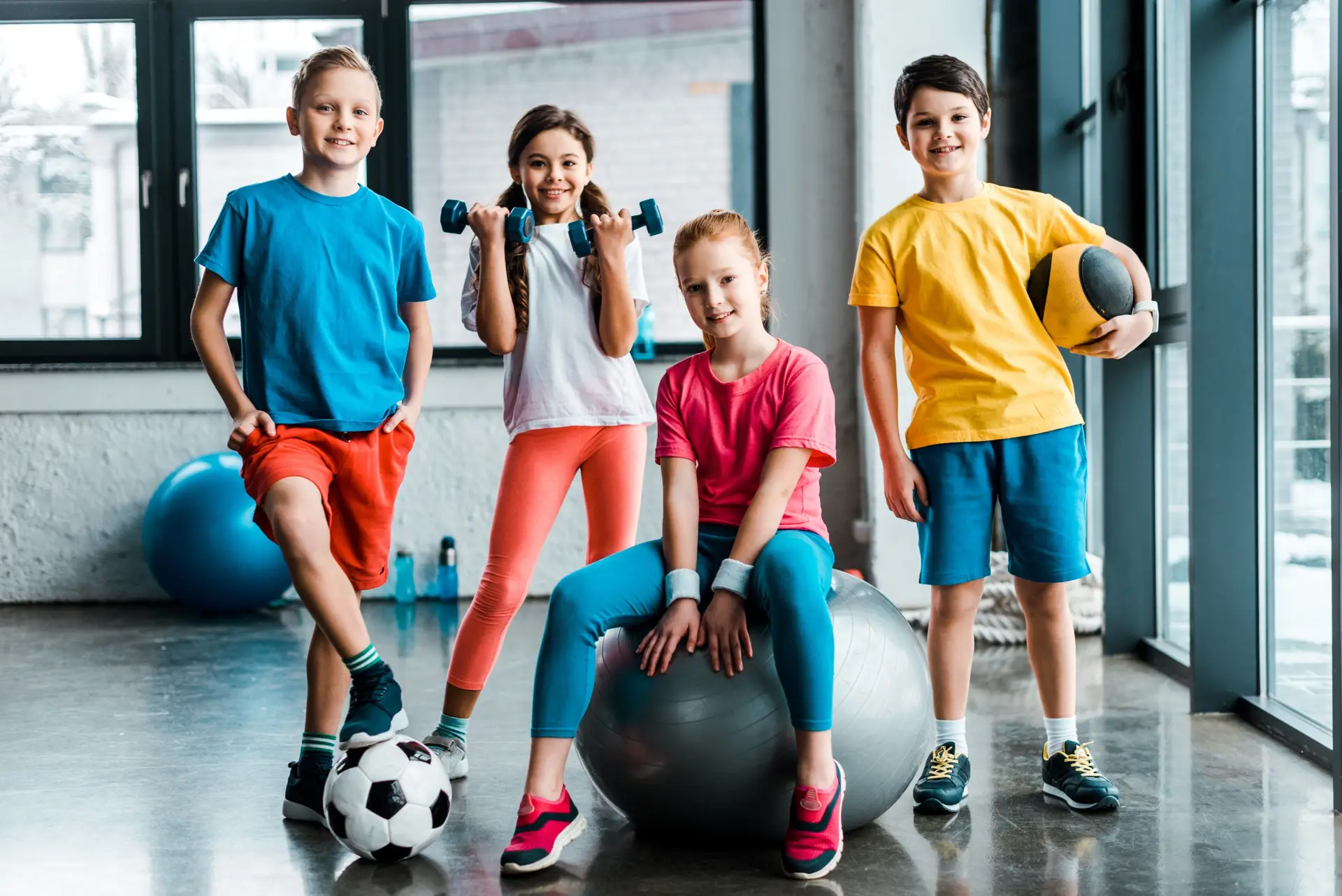 Four children in colorful athletic wear are in a gym. Smiling, one holds a soccer ball, another a basketball, while one lifts small weights and the last sits on an exercise ball. Surrounded by gym equipment and large windows, these kids embrace the Active Over Passive philosophy in their love for exercise.