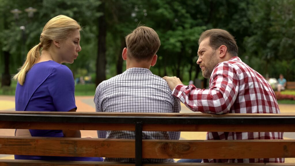 Three people sit on a park bench. A woman in a blue shirt talks to a man in a plaid shirt, who seems lost in thought as if contemplating suicide. Meanwhile, another man in a checked shirt sits between them. Trees and a walkway are visible in the background.