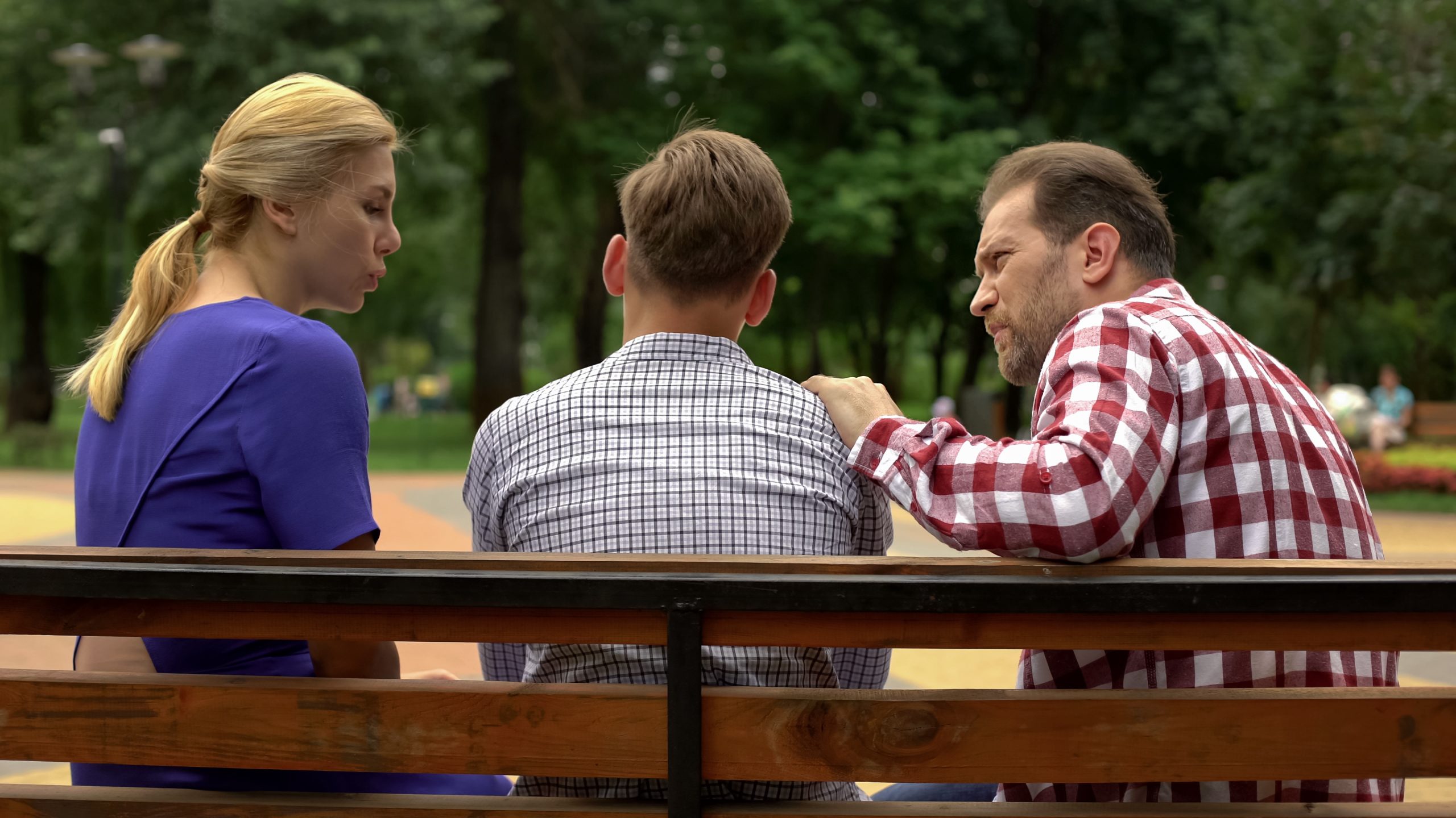 Three people sit on a park bench. A woman in a blue shirt talks to a man in a plaid shirt, who seems lost in thought as if contemplating suicide. Meanwhile, another man in a checked shirt sits between them. Trees and a walkway are visible in the background.