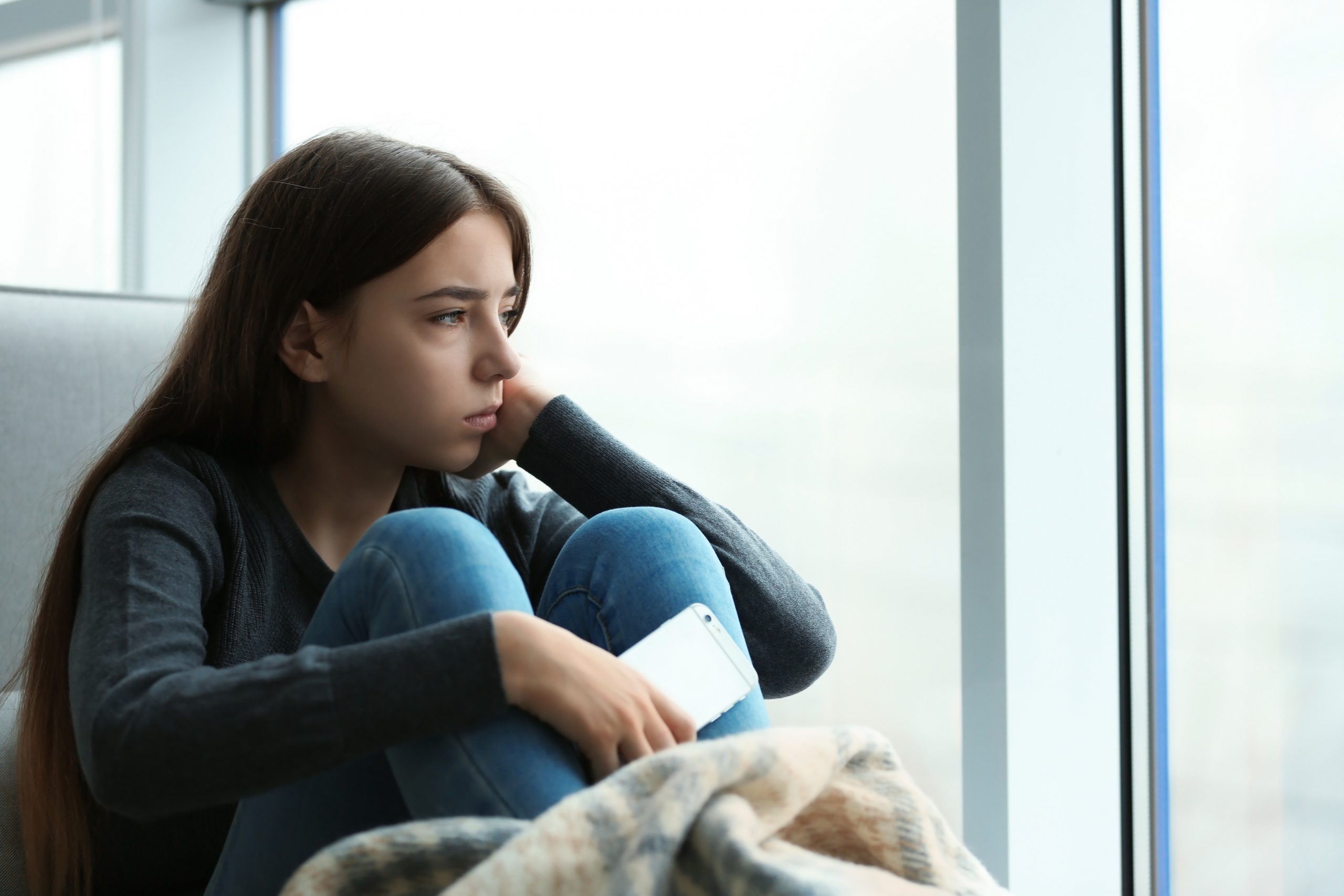 A young woman with long brown hair sits on a couch, gazing thoughtfully out a large window. Wrapped in a soft, patterned blanket and holding a smartphone, she appears deep in contemplation, perhaps reflecting on her mental health amid moments of crisis.