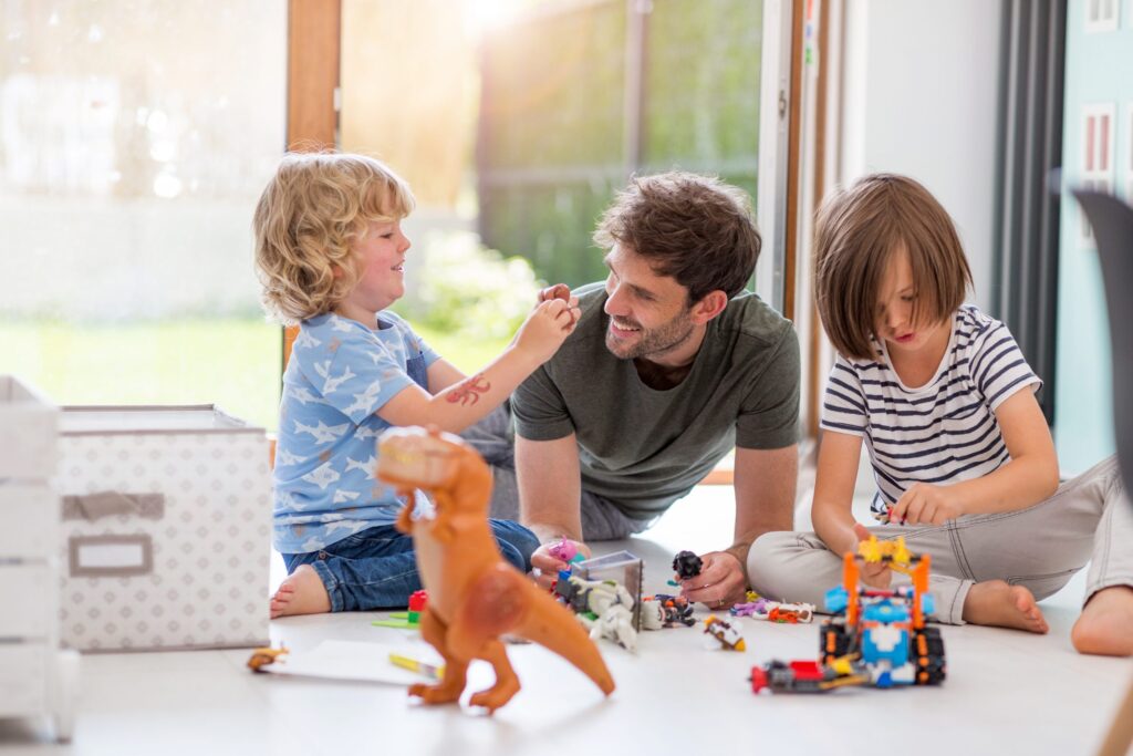 A man and two children are sitting safely on the floor playing with toys. The boy in blue smiles, holding a toy, while the girl in stripes focuses on assembling blocks—a scene of joyful play filled with cherished toys. A toy dinosaur stands in the foreground of their bright, cheerful room.