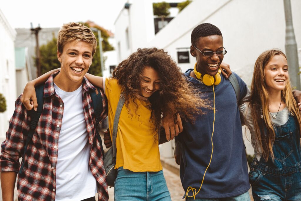 A group of four teenagers strolls outdoors, laughing and with arms around each other, momentarily free from the responsibilities of a teenager. They bask in the sunny day on a street lined with white buildings. One sports yellow headphones, while another rocks a plaid shirt.