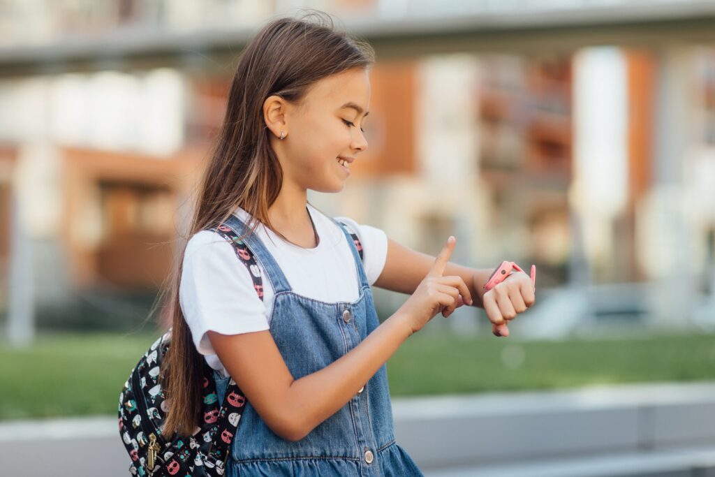 A young girl in a denim dress and backpack beams while using the best smartwatch phone for kids, pointing at its vibrant pink screen. The background is softly blurred, hinting at an outdoor urban setting where she explores her digital world with delight.