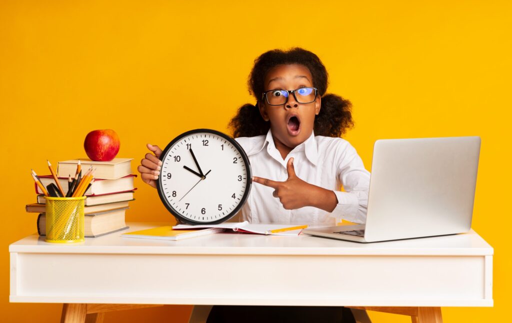 A girl with glasses and pigtails sits at a white desk against a yellow background, holding a large clock showing 10:10. She looks surprised, perhaps just realizing the importance of teaching teens time management. The desk has a laptop, books, a red apple, and a cup of pencils.