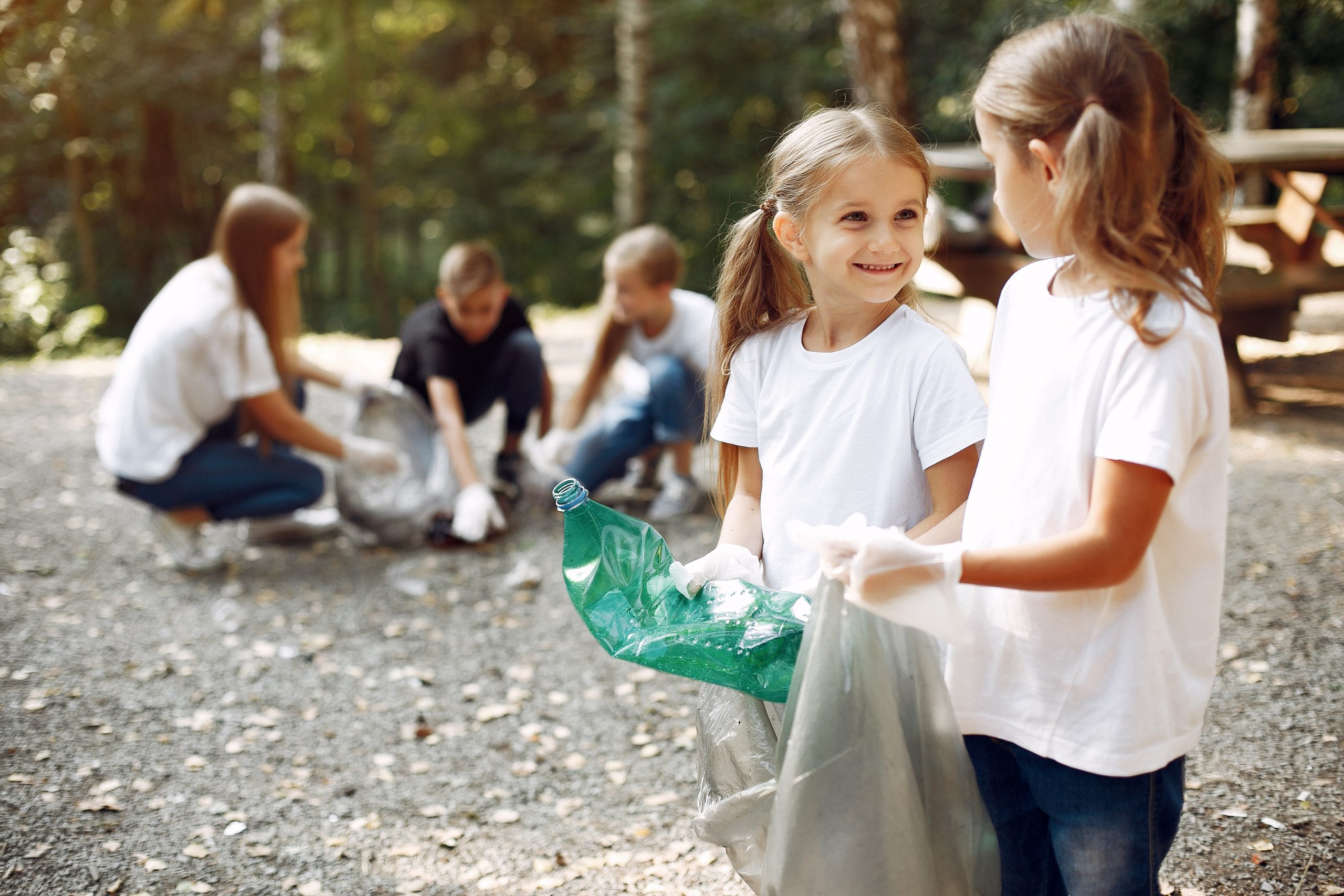 Two young girls in white shirts and gloves clean up a park area, holding a large green plastic bottle and a bag. In the spirit of Remembering September 11 with Service, three more people gather trash in the background. The scene is set in a wooded area with a picnic table nearby.