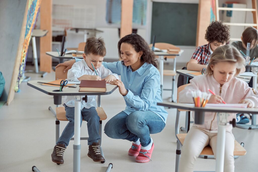 A teacher kneels next to a student, illustrating the importance of lifelong learning as she assists with his work. Other children sit at their desks, engrossed in their tasks. The room is bright and colorful, filled with diverse materials to foster curiosity and growth.