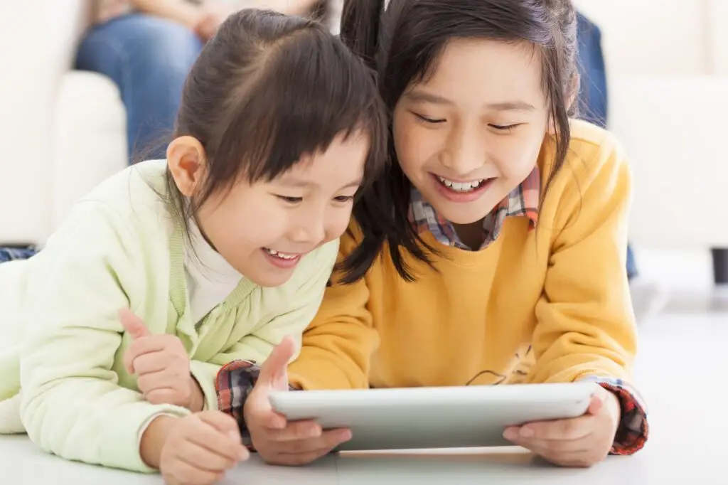 Two children are lying on the floor, smiling and looking at a tablet. The child on the left is wearing a light green sweater, while the one on the right sports yellow. They seem to be enjoying their content—a sweet glimpse into when to introduce a tablet to a child.