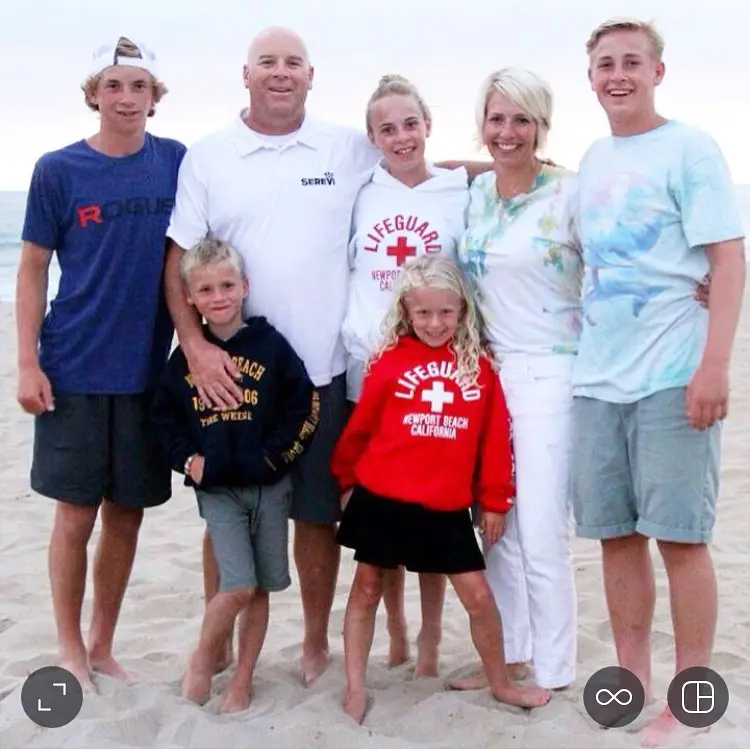 A family of seven poses on a sandy beach. The group includes two adults and five children, some of whom wear casual clothing, such as lifeguard hoodies supporting suicide awareness. They are smiling, and the sky is overcast.