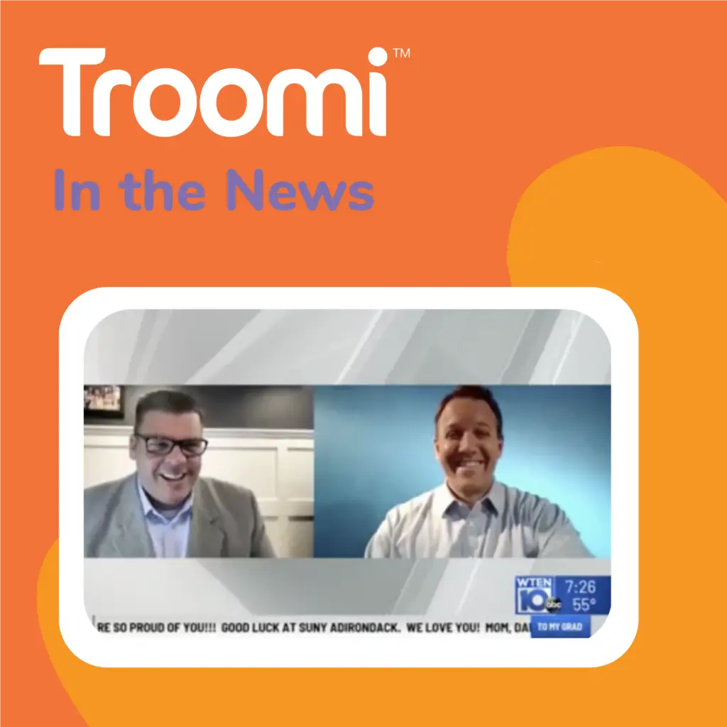 Two men in suits are on a video call, smiling. The screen features Troomi In the News on an orange background, highlighting their innovative phone that adjusts to your family’s needs. Below, a news ticker scrolls with congratulatory messages, celebrating this technological advancement.