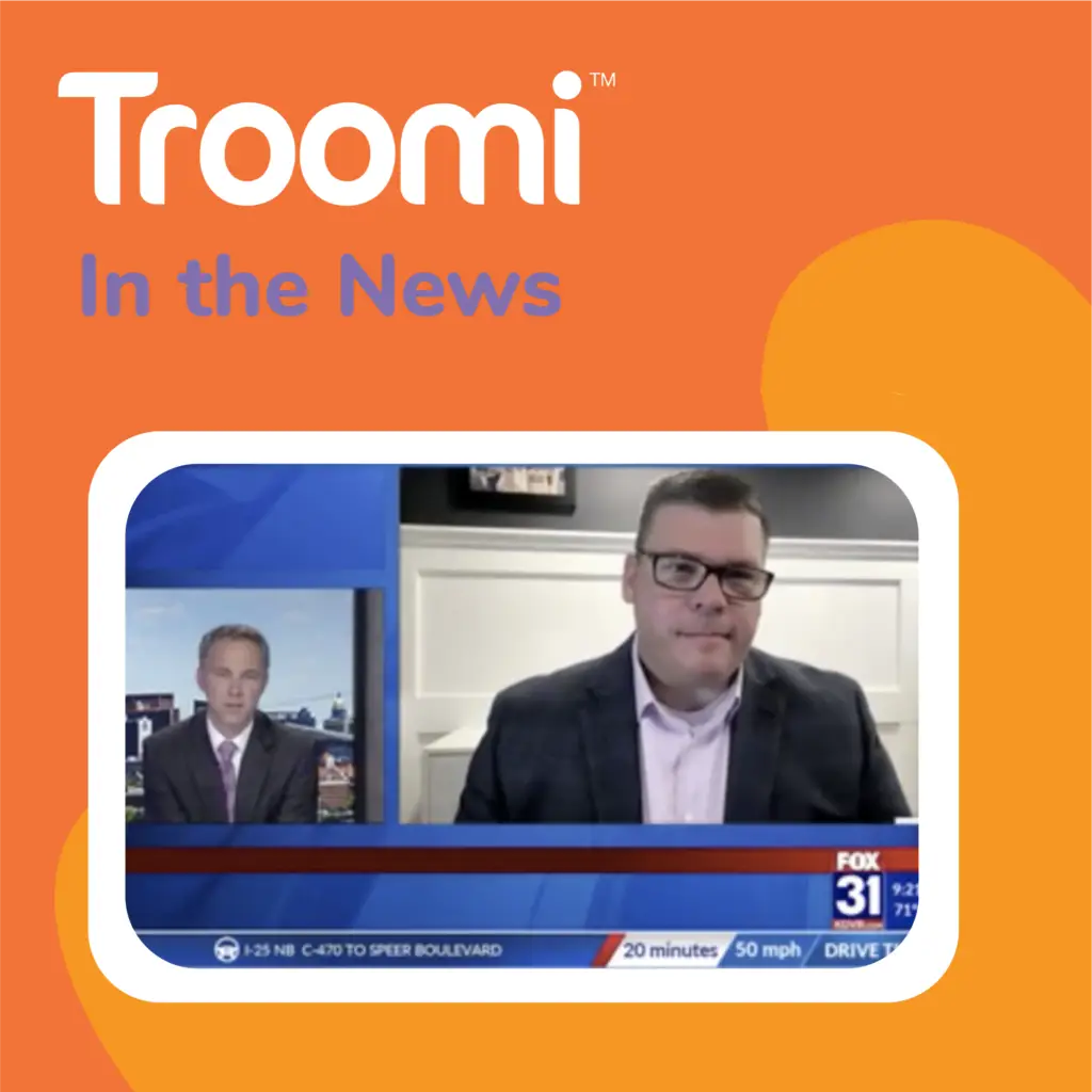 A news broadcast features two men. One is in a studio, while the other appears via video call discussing a phone that adjusts to your family's needs. The screen is labeled Fox 31 with traffic updates displayed, and the background is branded with Troomi In the News text.