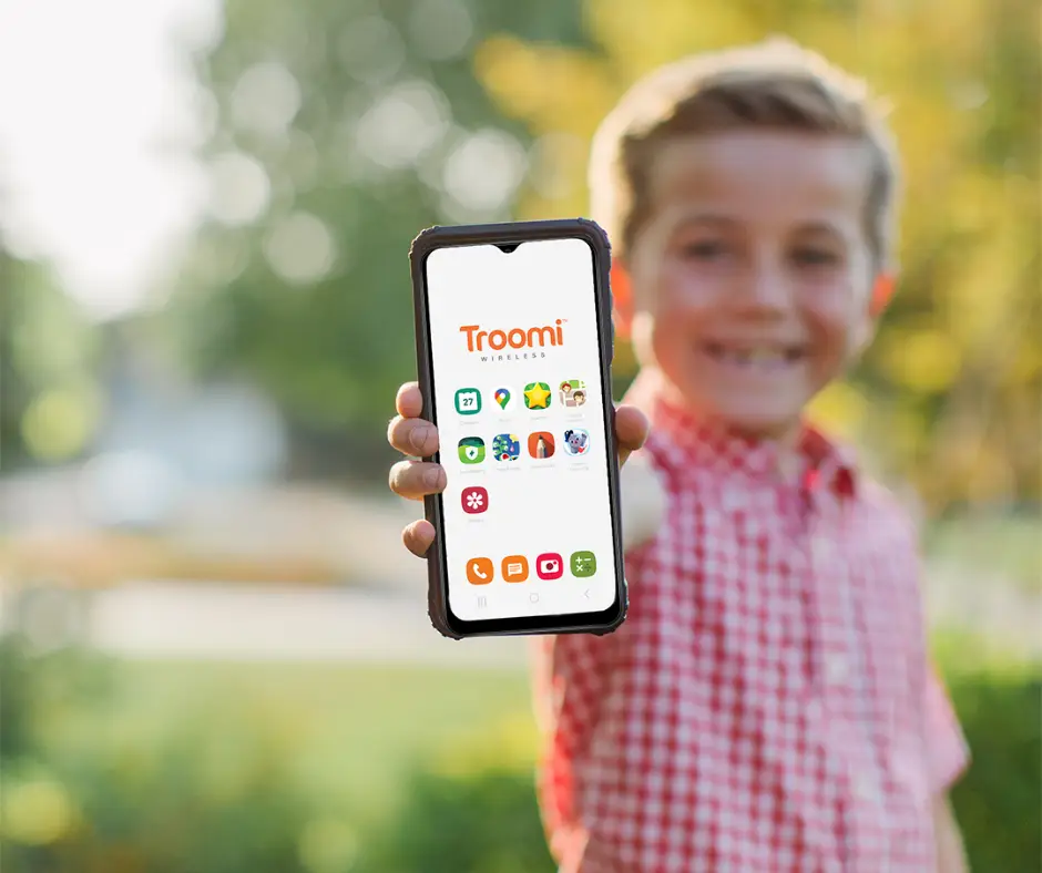 A smiling boy in a red checkered shirt proudly holds the best cell phone for a kid, displaying the Troomi Wireless home screen with various app icons. In the background, a lush outdoor setting adds to his joy and sense of adventure.