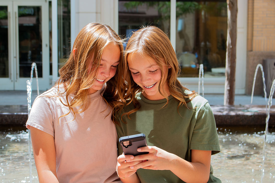 Two smiling young girls sit by a fountain, sharing laughter as they look at a smartphone. One wears a pink shirt and the other green, perhaps discussing internal research from Facebook that uncovers Instagram's pitfalls while enjoying their moment of connection.