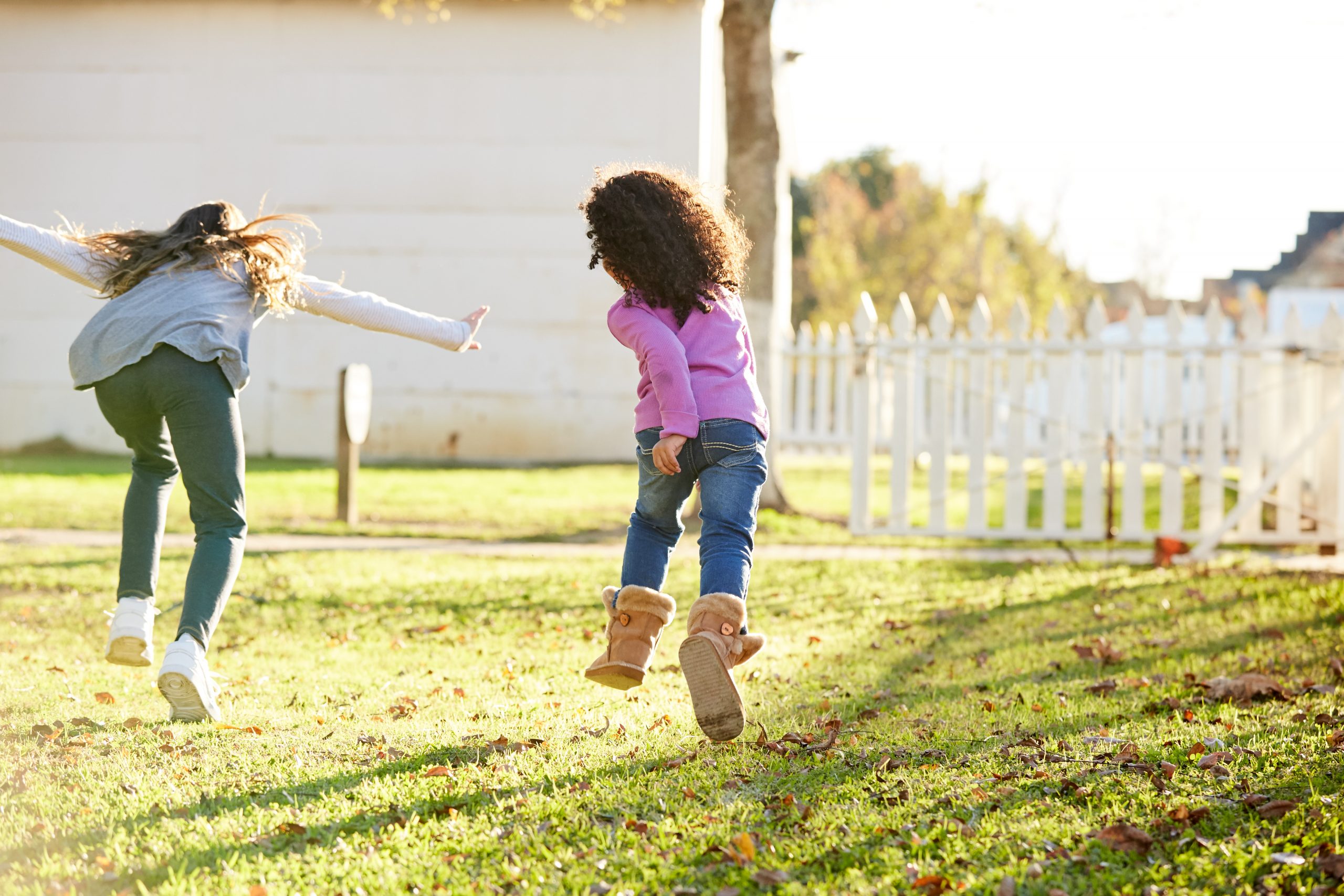 Two children run joyfully across a sunlit grassy yard, showcasing how to enjoy your backyard. One wears a gray sweater and jeans, the other a purple top and jeans with boots. A white picket fence and trees are in the background, with a house partially visible.