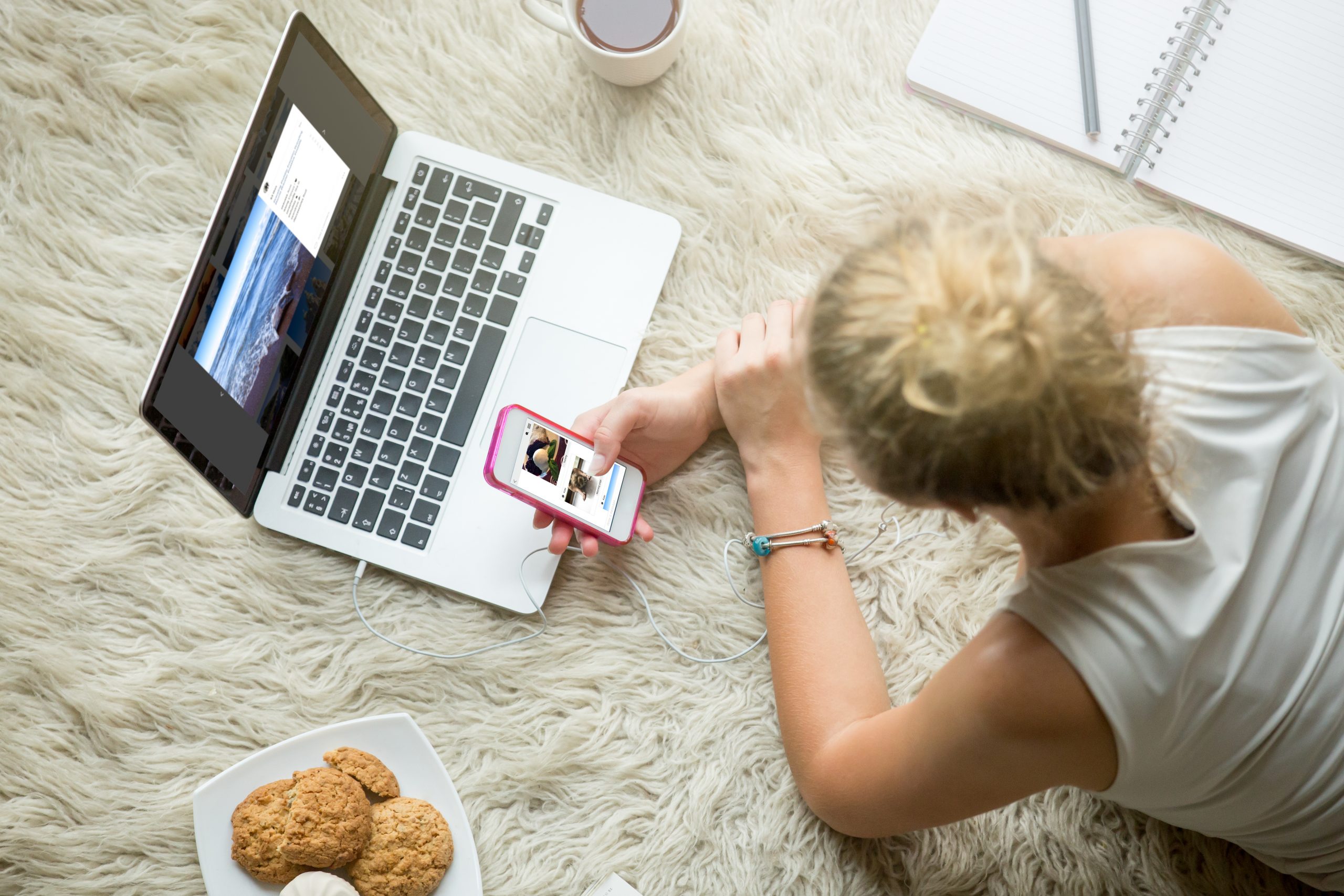 A person with blond hair lies on a fuzzy rug, scrolling through their smartphone while an open laptop sits nearby. As they sip coffee and nibble cookies, they wonder: is Instagram safe for kids? The cozy setup includes a notebook, completing the scene's inviting vibe.