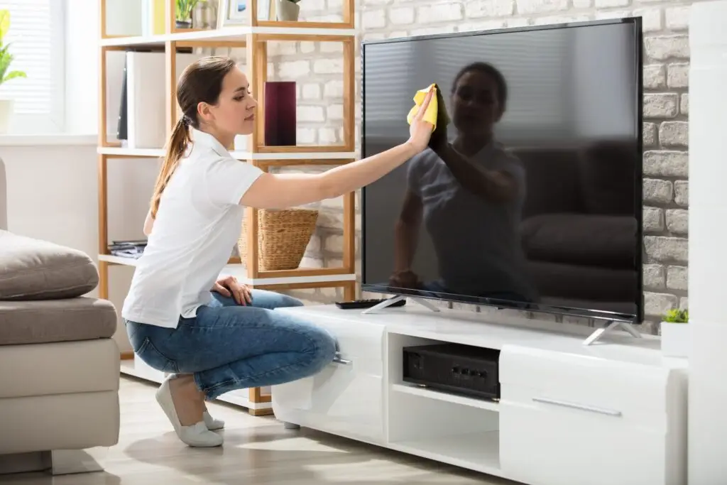 A woman in a white shirt and blue jeans demonstrates how to safely clean TV screens as she kneels, using a yellow cloth on a flat-screen. She's in a living room complete with a brick wall, wooden shelves, and a gray sofa.