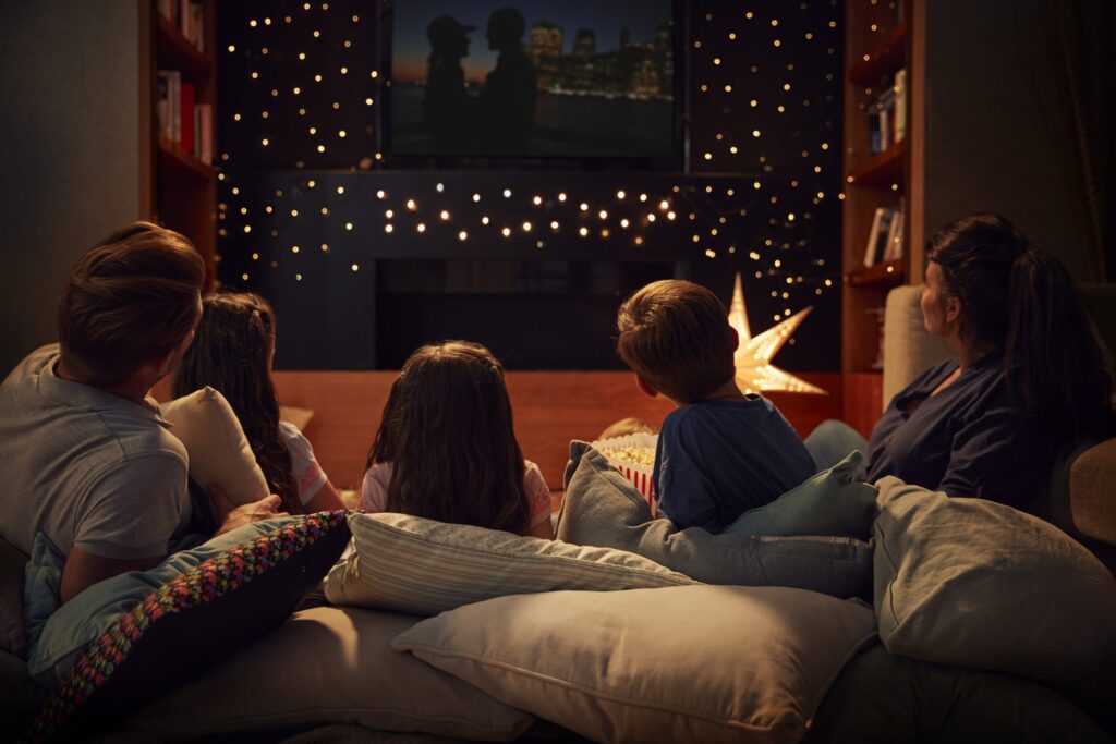 A family of four enjoys one of eight Halloween-themed movies in their cozy living room. They sit on cushioned floor seating, surrounded by soft pillows. A bowl of popcorn is nearby, and a star-shaped light decoration adds a warm ambiance.